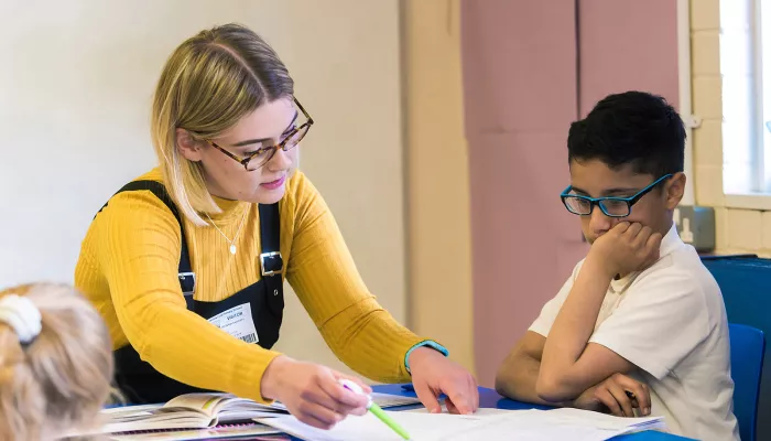 A tutor in a yellow jumper wearing dungarees is explaining a document to a young boy wearing blue glasses.