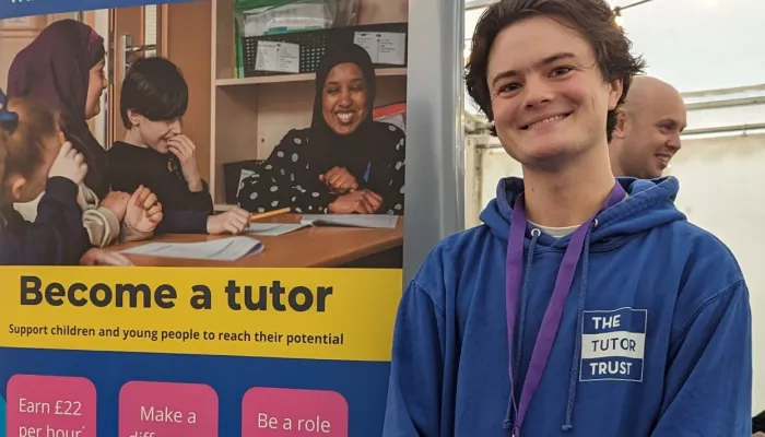 A colleague from the Recruitment Team drums up support at a Jobs Fair at the University of Liverpool. He stands in front of a banner giving details of the role.  He is wearing a blue branded Tutor Trust hoodie.