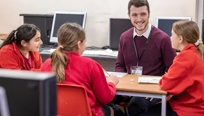 A male tutor smiles at three pupils, who all seem to be enjoying the tuition session. The pupils are wearing red jumpers.