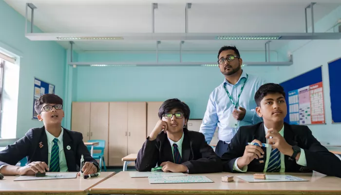 Three secondary school-aged male pupils wearing school uniform are sat at a desk looking up at a screen that is behind the camera. Behind them stands a male tutor wearing a blue shirt and glasses. They are in a classroom with a pale blue back wall.