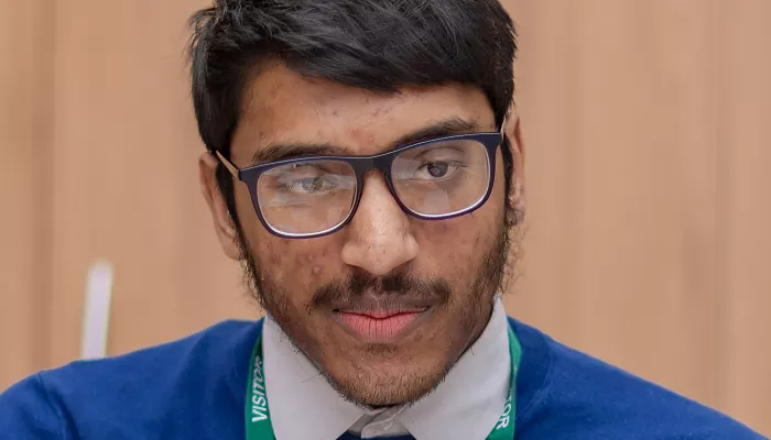 Farhan is wearing a dark blue round-necked jumper with a shirt underneath. He is wearing glasses.  He is looking at one of his tutees.