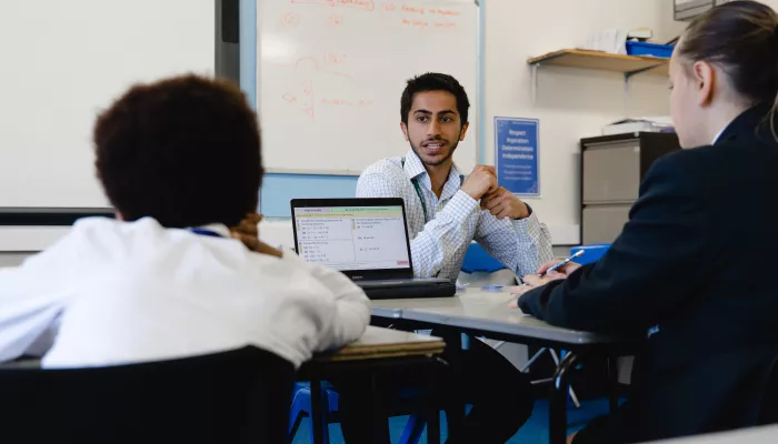 In the centre of the image a male tutor wearing a light blue shirt is sat at a desk facing the camera. He is looking at two pupils who have their backs to the camera. They are sat at desks looking at laptop computers.