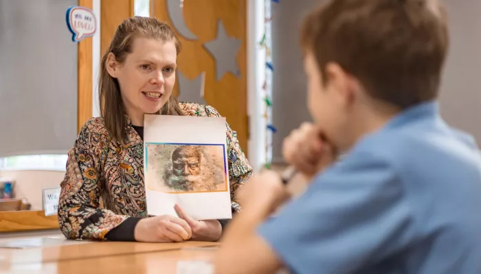 A tutor holds up an image and is speaking to a pupil