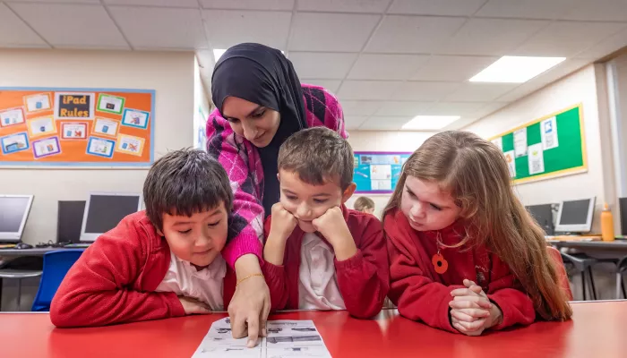 A female tutor wearing a black hijab and pink sweater is stood behind three primary-aged pupils who are sat around a table. They are wearing red and white school uniforms. The tutor is pointing out a concept on a worksheet. The children are concentrating.