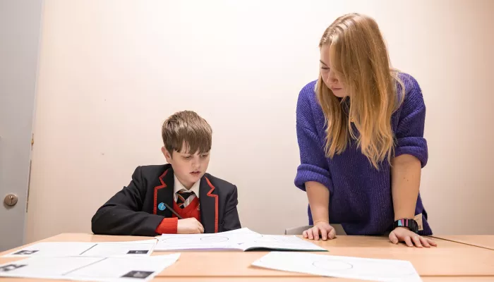 A boy in a black school uniform is sat at a desk reading a text. To his right a female tutor with long blonde hair and a purple jumper is stood up helping him with his work. 