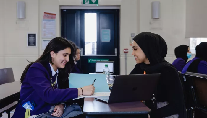 A secondary aged female pupil with long dark hair, wearing a purple blazer, is sat at a desk writing. An open laptop is on the desk. To the right of the desk is sat a young female tutor wearing a black jumper and black hijab.