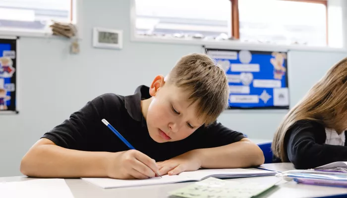 A primary school-aged boy wearing a black t-shirt is sat at a desk and is writing with a pencil in an exercise book.