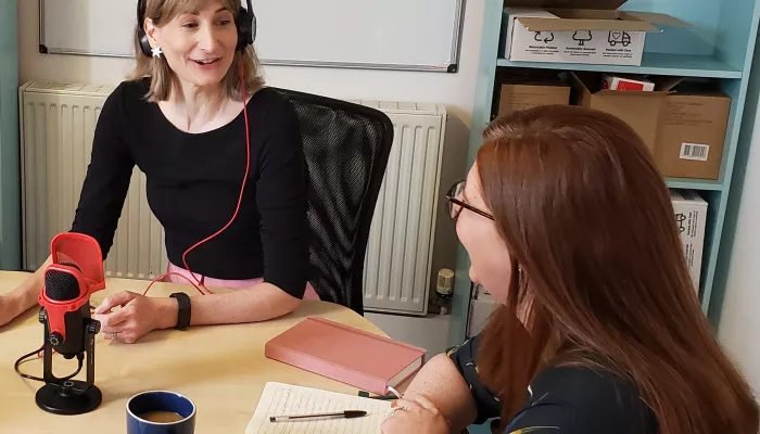 A room has been set up as a recording studio with microphone in the centre of the table.  The interviewer, wearing headphones, is smiling at her guest.