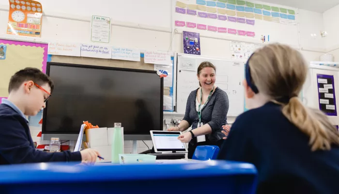 A female tutor is standing in front of a whiteboard, next to a large TV that is turned off. She is teaching two primary-aged pupils, whose faces are not visible. The tutor is smiling.