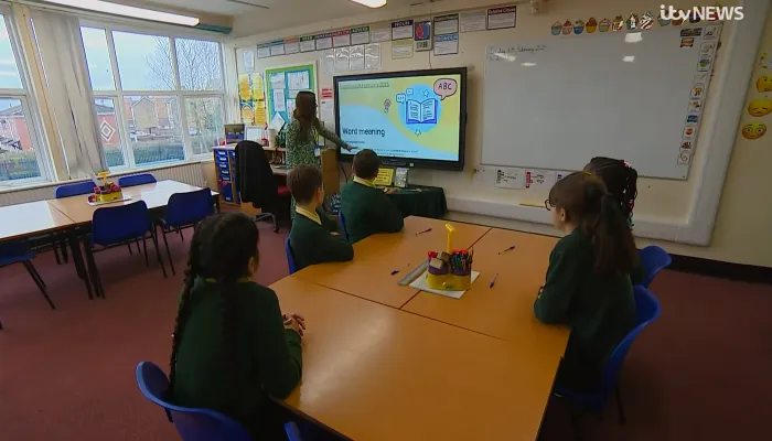 a teacher in a green dress in front of a whiteboard and some primary school pupils