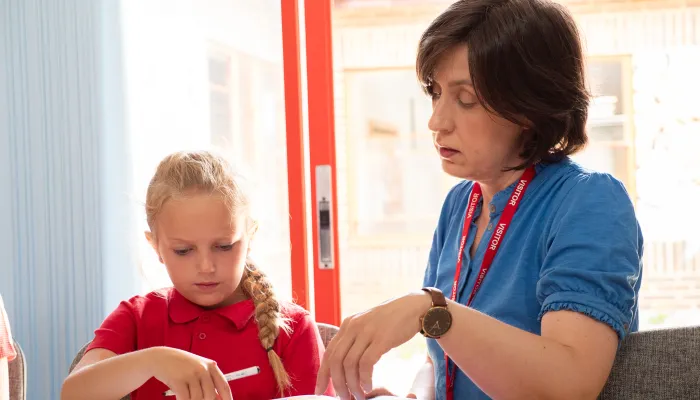 A female tutor with brown hair and a blue top sitting at a table with a primary school child in a red polo shirt.