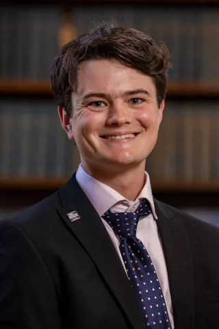 Theo, a former tutor, now ensures more young people, like him, can take up the opportunity. He is wearing a white shirt, with a tie and jacket.