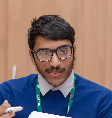 Farhan is wearing a dark blue round-necked jumper with a shirt underneath. He is waring glasses.  He is looking at one of his tutees.