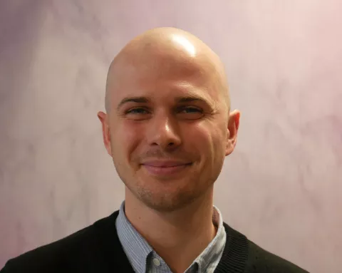 Ed Thompson is a recruitment coordinator passionate about the role of tutoring for young people. He is smiling, wearing a grey open necked shirt.