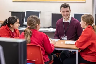 A male tutor smiles at three pupils, who all seem to be enjoying the tuition session. The pupils are wearing red jumpers.