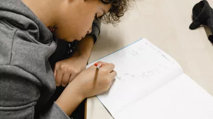 A close up photo of a secondary school-aged male pupil writing in an exercise book. He is sat at a desk wearing a grey jumper and has brown hair.