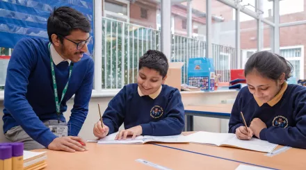 A male tutor with black hair and glasses, wearing a blue jumper, is leaning over a desk. Two primary aged pupils, a boy and a girl, wearing dark blue school jumpers are holding pencils and writing in exercise books. Everyone is smiling.