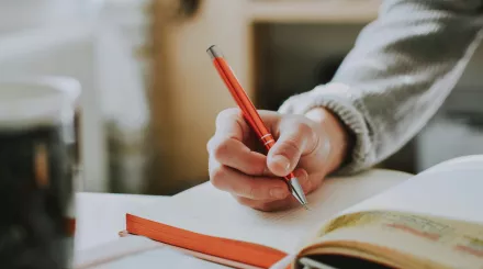 A close up shot of a person's hand above a notebook. The person is wearing a light grey jumper and holding a dark orange pen.