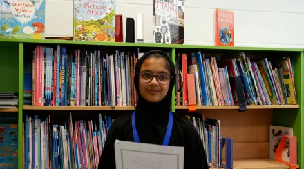 Umme is a primary school pupil and winner of the Speak Up North competition. She wears glasses and smiles to camera while holding and reading her poem.