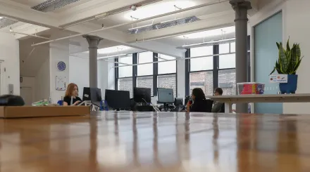 Tutor Trust's new Manchester office. A wooden worksurface is in the foreground, whilst grey pillars and computer workstations sit further back