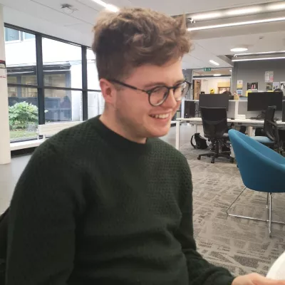 James supports the digital growth of Tutor Trust and focuses on sharing the story of our impact. He has brown hair and is wearing glasses. He is wearing a dark blue top.