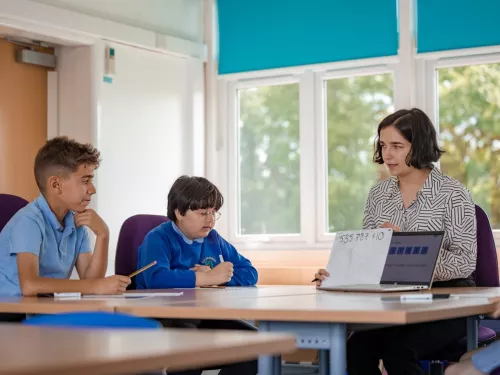 two primary school boys wearing blue jumpers, receiving tutoring from a female tutor with short dark hair