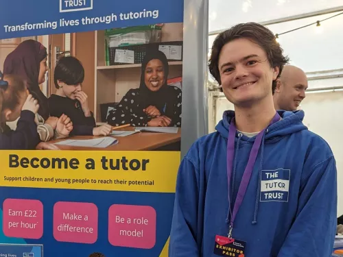 A colleague from the Recruitment Team drums up support at a Jobs Fair at the University of Liverpool. He stands in front of a banner giving details of the role.  He is wearing a blue branded Tutor Trust hoodie.