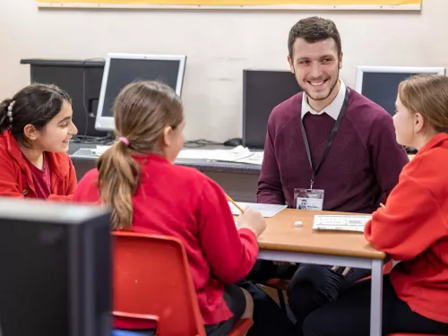 A male tutor smiles at three pupils, who all seem to be enjoying the tuition session. The pupils are wearing red jumpers.