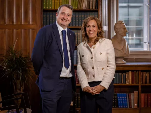 A man and a woman, standing in front of a bookcase, smile for the camera.