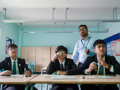 Three secondary school-aged male pupils wearing school uniform are sat at a desk looking up at a screen that is behind the camera. Behind them stands a male tutor wearing a blue shirt and glasses. They are in a classroom with a pale blue back wall.