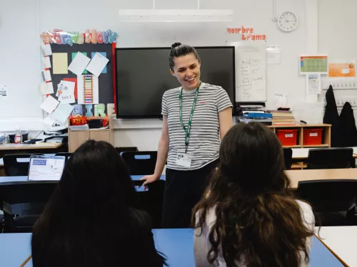 A young female tutor with dark hair, tied in a bun, wearing a striped t-shirt, is standing talking to two secondary-aged female pupils who are sat at a desk. The backs of the pupils' heads are facing the camera. Both pupils have long dark hair.