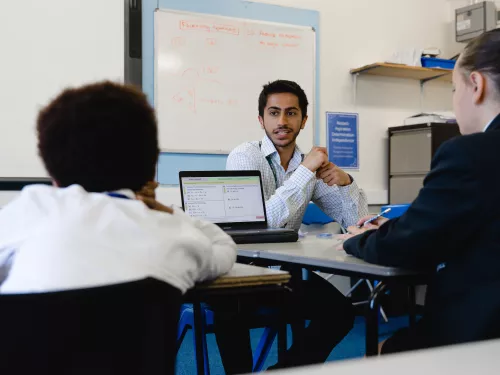 In the centre of the image a male tutor wearing a light blue shirt is sat at a desk facing the camera. He is looking at two pupils who have their backs to the camera. They are sat at desks looking at laptop computers.