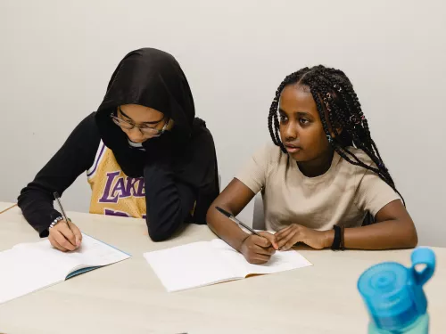 Two secondary-school age female pupils are sat at a desk. The one on the left is wearing a long black top, LA Lakers t-shirt and black hijab, the one on the right has long black braided hair and is wearing a beige t-shirt. They are writing in exercise books.