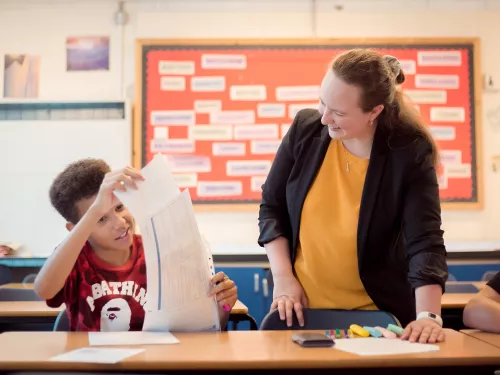 On the right of the image a female tutor wearing a yellow top and black blazer is standing at a desk. Sat next to her at the desk is a male pupil wearing a red t-shirt. He is holding up a large sheet of paper. The tutor and the pupil are both smiling.
