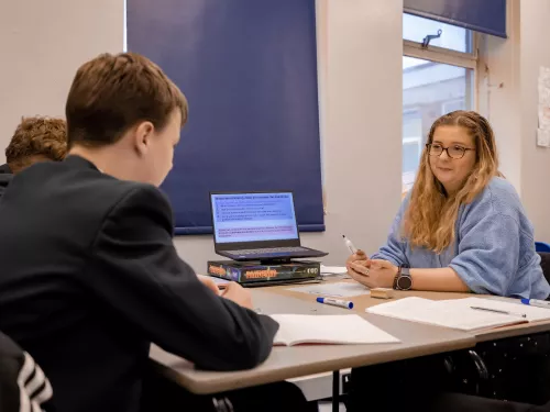 To the left of the photo is a male secondary school-aged pupil wearing a dark blazer. He is turned away from the camera and sat at a desk. To the right is a female tutor with long blonde hair wearing a blue sweater. On the middle of the desk is an open laptop.
