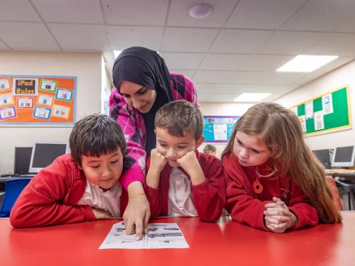 A female tutor wearing a black hijab and pink jumper is standing above three primary aged pupils wearing red school uniforms. She is pointing at something on a worksheet. The children are concentraing.