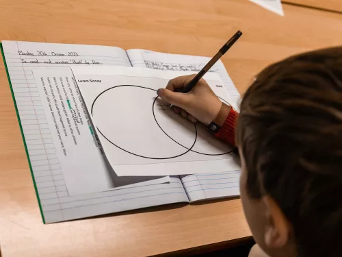 A close up photo of a school exercise book on a brown wooden desk. The exercise book is open to a Venn diagram and a male secondary student is using a pen to complete the Venn diagram. Only the back of his head is visible in the shot.