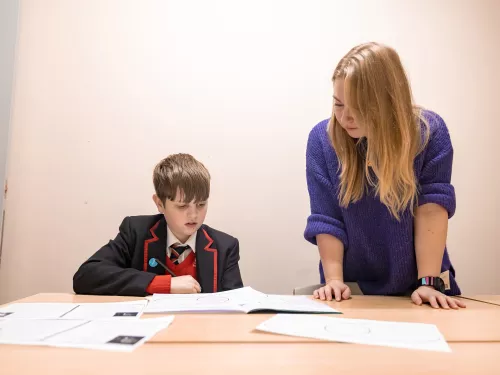 A boy wearing a school uniform of a black blazer and red jumper is reading through a text. To his right a female tutor with long blonde hair wearing a purple jumper is standing. She is supporting him with his work.