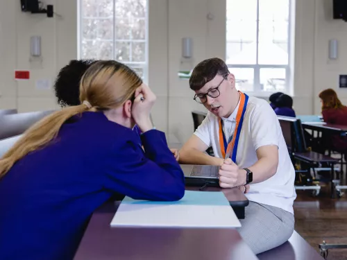 To the left of the picture two secondary school-aged female pupils wearing purple blazers are sat at a desk with their backs to the camera. On the other side of a desk a young male tutor wearing a white t-shirt and glasses is sat. He is looking down at a laptop and explaining a concept to the pupils.