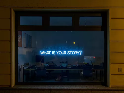 A full-width shop window. In the window is a small blue neon sign that says 'what is your story?' in capital letters.