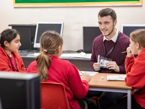 A male tutor wearing a burgundy jumper is sat tutoring a group of primary school-aged pupils wearing bright red school uniforms.