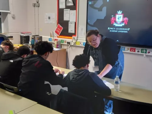 A female tutor stands in front of a screen, speaking to two male pupils sitting a desk and wearing hoodies 