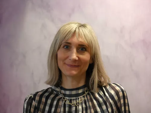 A blonde woman in a checked blouse stands in front of a lilac background