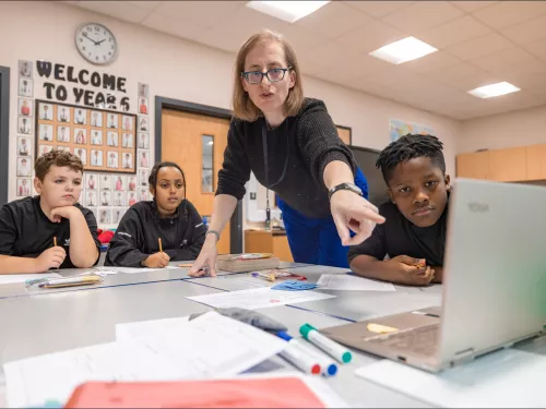 A female tutor sands and points at a screen which three seated children also look at