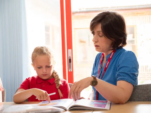 A female tutor with brown hair and a blue top sitting at a table with a primary school child in a red polo shirt.